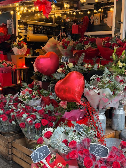 A vibrant display of fresh floral arrangements and bouquets featuring red roses, white flowers, and eucalyptus foliage, arranged in wicker baskets, gift boxes, and wrapped in decorative pink and red p
