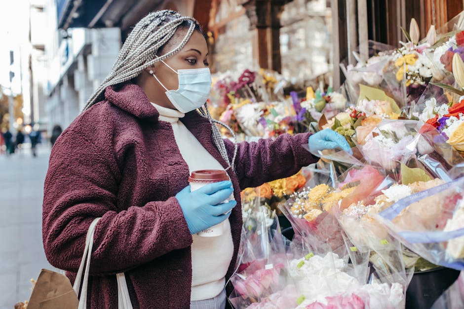A woman wearing a light blue face mask and blue gloves is standing outdoors at a flower market on a city street. She is dressed in a burgundy fuzzy jacket over a white sweater, with braided hair. She