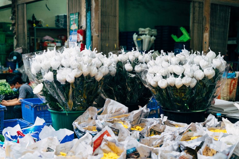 A display of white roses wrapped in clear plastic, arranged in large black and teal buckets on a market stall at South Hackney Florist. The roses have fresh, slightly closed buds with green stems and